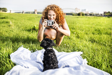 Deutschland, Köln, Porträt einer lächelnden jungen Frau auf einer Wiese, die ihren Hund fotografiert - FMKF04465