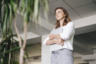 Businesswoman standing in office, holding laptop - KNSF02718