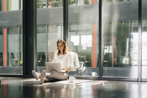 Businesswoman sitting on ground in empty office, using laptop - KNSF02723