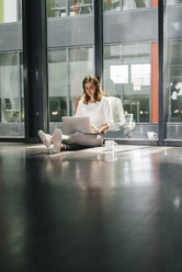 Businesswoman sitting on ground in empty office, using laptop - KNSF02724