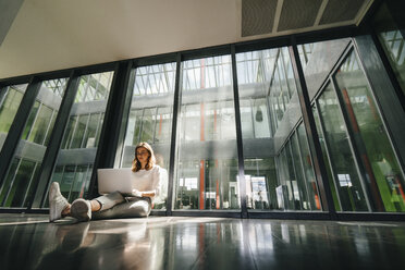 Businesswoman sitting on ground in empty office, using laptop - KNSF02732