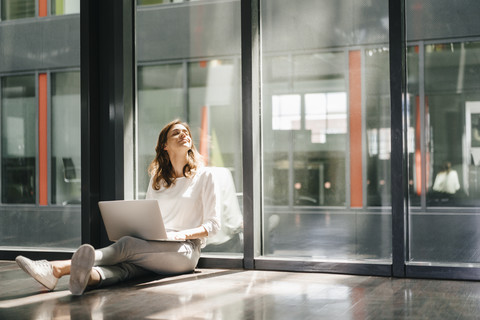 Businesswoman sitting on ground in empty office, using laptop stock photo