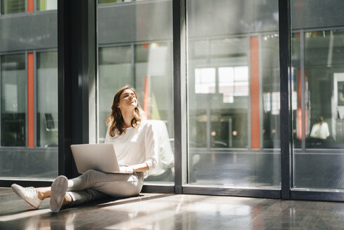 Businesswoman sitting on ground in empty office, using laptop - KNSF02734