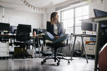 Businesswoman sitting cross-legged on an office chair - KNSF02751