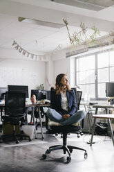 Businesswoman sitting cross-legged on an office chair - KNSF02752