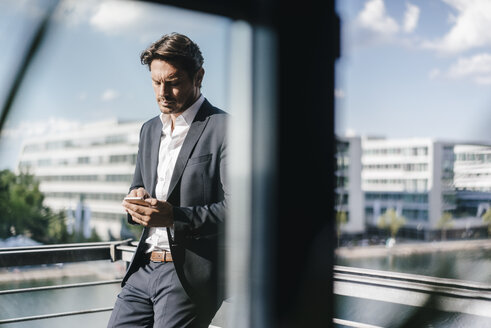 Businessman standing on balcony, holding smartphone - KNSF02774