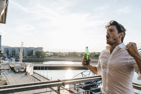 Happy businessman standing on balcony, drinking beer - KNSF02815