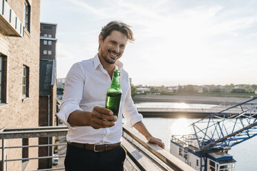 Happy businessman standing on balcony, drinking beer - KNSF02816