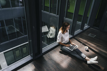 Businesswoman sitting on ground in empty office, using laptop - KNSF02825