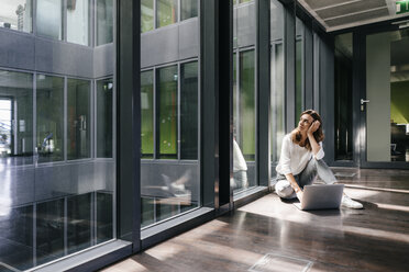 Sad businesswoman sitting on ground in empty office, using laptop - KNSF02826