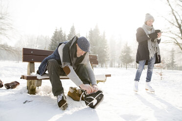 Senior man sitting on bench in winter landscape putting on ice skates - HAPF02148
