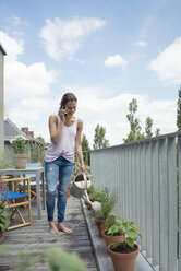 Woman on cell phone on balcony watering plants - JOSF01553