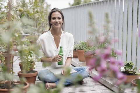 Lächelnde Frau mit Flasche und Laptop entspannt auf dem Balkon, lizenzfreies Stockfoto