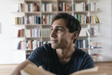 Lächelnder junger Mann mit Buch zu Hause mit Blick zur Seite - JOSF01585