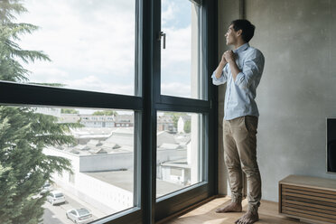 Young man buttoning his shirt looking out of window - JOSF01597