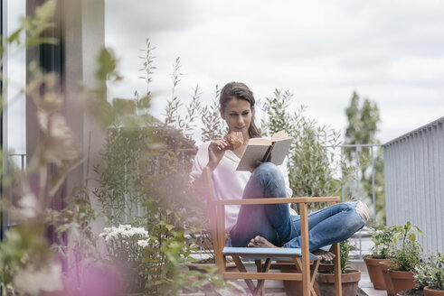 Woman eating croissant and reading book on balcony - JOSF01637