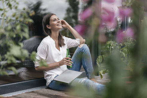 Glückliche Frau mit Laptop und Flasche auf dem Balkon sitzend - JOSF01644