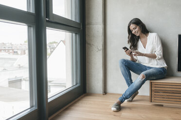 Lächelnde Frau, die sich hinsetzt und auf ihr Mobiltelefon schaut - JOSF01651