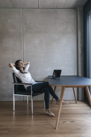 Frau sitzt mit Laptop am Tisch und lehnt sich zurück, lizenzfreies Stockfoto