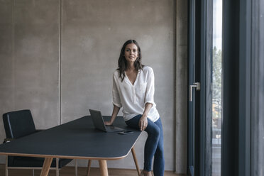 Portrait of smiling woman sitting on table with laptop and cell phone - JOSF01663