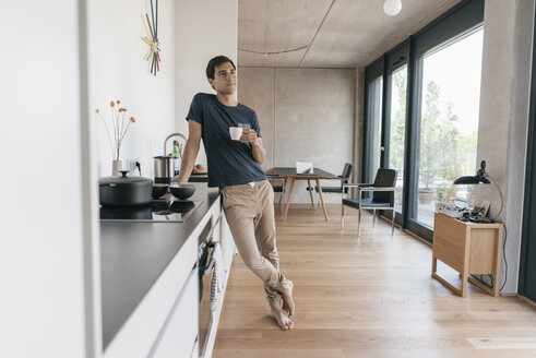 Young man holding cup of coffee in kitchen at home - JOSF01673