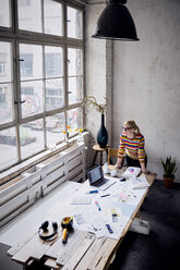 Frau steht am Schreibtisch in einem Loft und schaut durch ein Fenster - RBF05954