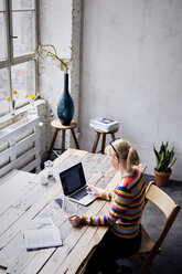 Frau sitzt am Schreibtisch in einem Loft und benutzt Laptop und Tablet - RBF05955