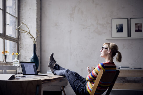 Smiling woman sitting with feet up at desk in a loft looking through window - RBF05956