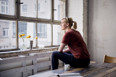 Woman sitting on desk in loft looking through window - RBF05957