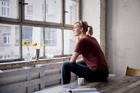 Frau sitzt auf einem Schreibtisch in einem Loft und schaut durch ein Fenster - RBF05957