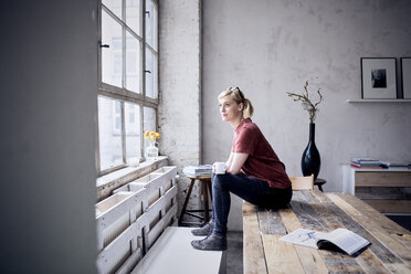 Smiling woman with coffee mug sitting on desk in loft looking through window - RBF05958