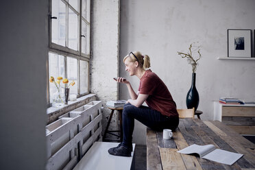 Woman sitting on desk in loft using cell phone - RBF05959