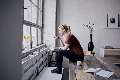 Frau sitzt auf einem Schreibtisch im Loft und benutzt ein Mobiltelefon - RBF05959