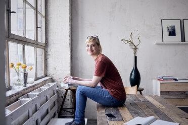 Porträt einer lächelnden Frau mit Kaffeetasse auf dem Schreibtisch in einem Loft - RBF05965