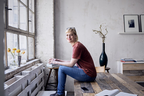Portrait of smiling woman with coffee mug sitting on desk in loft - RBF05965