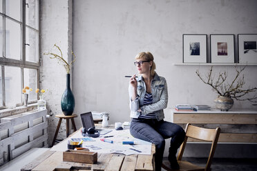 Frau sitzt auf einem Schreibtisch in einem Loft und schaut durch ein Fenster - RBF05968