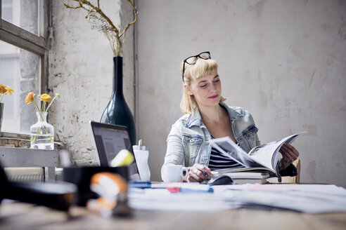 Porträt einer lächelnden Frau, die an einem Schreibtisch in einem Loft sitzt und in einem Buch blättert - RBF05975