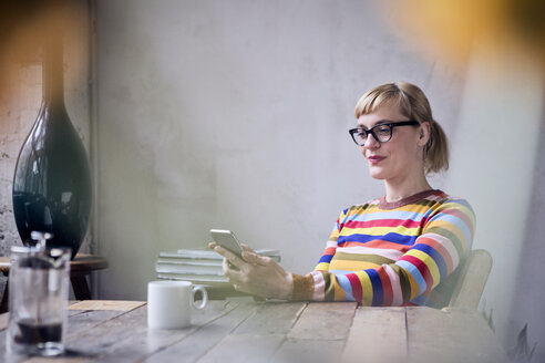 Porträt einer lächelnden Frau, die mit einer Tasse Kaffee am Tisch sitzt und auf ihr Mobiltelefon schaut - RBF05987