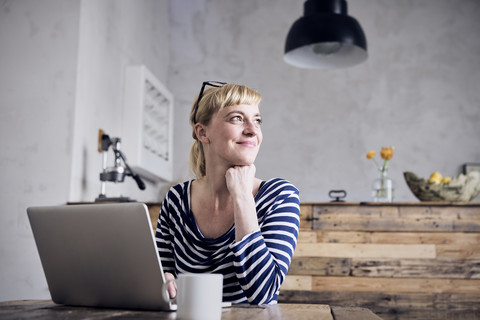 Porträt einer lächelnden Frau, die mit Laptop und Kaffeetasse am Tisch sitzt, lizenzfreies Stockfoto