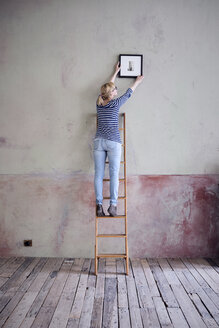 back view of woman on ladder hanging up picture frame in an unrenovated room of a loft - RBF06012