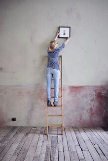 rückenansicht einer Frau auf einer Leiter, die einen Bilderrahmen in einem unrenovierten Raum eines Dachbodens aufhängt - RBF06012