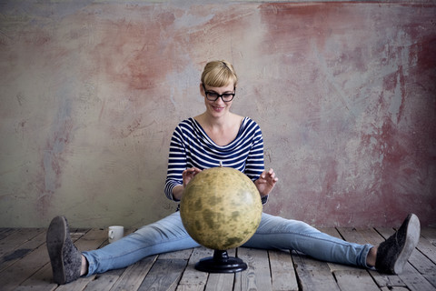 Smiling woman sitting on wooden floor in an unrenovated room looking at globe stock photo