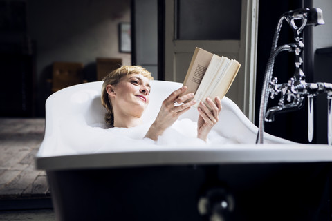 Portrait of relaxed woman taking bubble bath in a loft reading a book stock photo