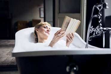 Porträt einer entspannten Frau, die in einem Loft ein Schaumbad nimmt und ein Buch liest - RBF06020