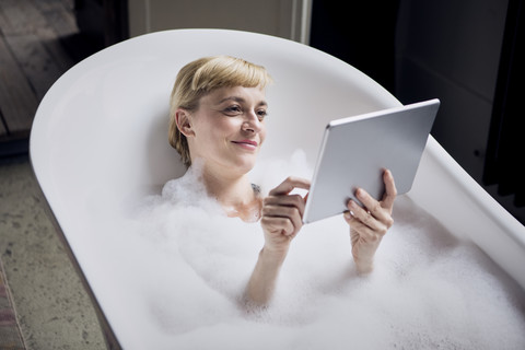 Porträt einer entspannten Frau, die ein Schaumbad nimmt und eine Tablet benutzt, lizenzfreies Stockfoto