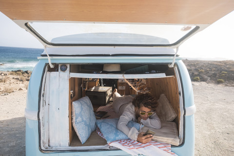 Spanien, Teneriffa, Frau mit Mobiltelefon in einem am Meer geparkten Lieferwagen liegend, lizenzfreies Stockfoto