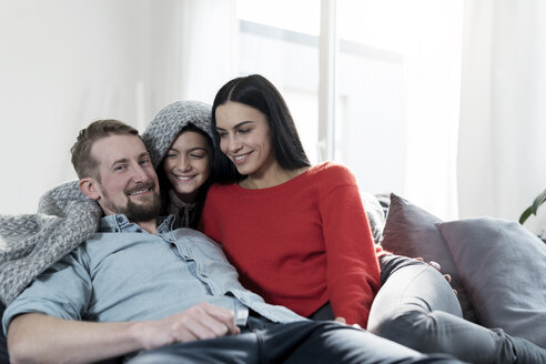 Eltern und Tochter kuscheln unter einer Decke auf dem Sofa im Wohnzimmer - SBOF00624