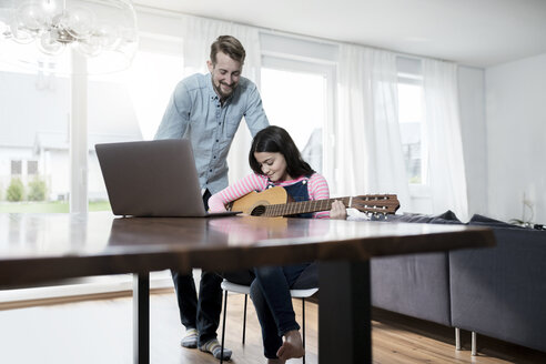 Vater lächelt Tochter an, die vor einem Laptop Gitarre spielt - SBOF00642
