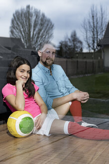 Mädchen im Fußball-Outfit sitzt neben ihrem Vater auf dem Boden im Wohnzimmer und schaut aus dem Fenster - SBOF00650