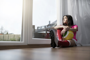 Mädchen im Fußball-Outfit sitzt auf dem Boden im Wohnzimmer und schaut aus dem Fenster - SBOF00653